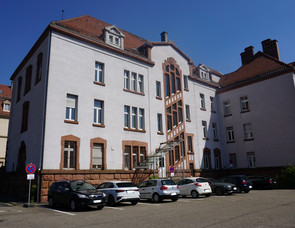 Foto Lernort Ansicht Parkplatz; Bildunterschrift: Auf der Rückseite des Gebäudes wird sich der Eingang zum Lernort befinden, dieser Eingang wurde früher auch für die Inhaftierten genutzt, wenn diese in die Zellen gebracht, oder zu Verhören oder zum Exerzieren im Hof heraufgeholt wurden. Diese Ansicht des Gebäudes findet sich auch im Logo-Design wieder, welches das Historische mit dem Heutigen verbindet.]